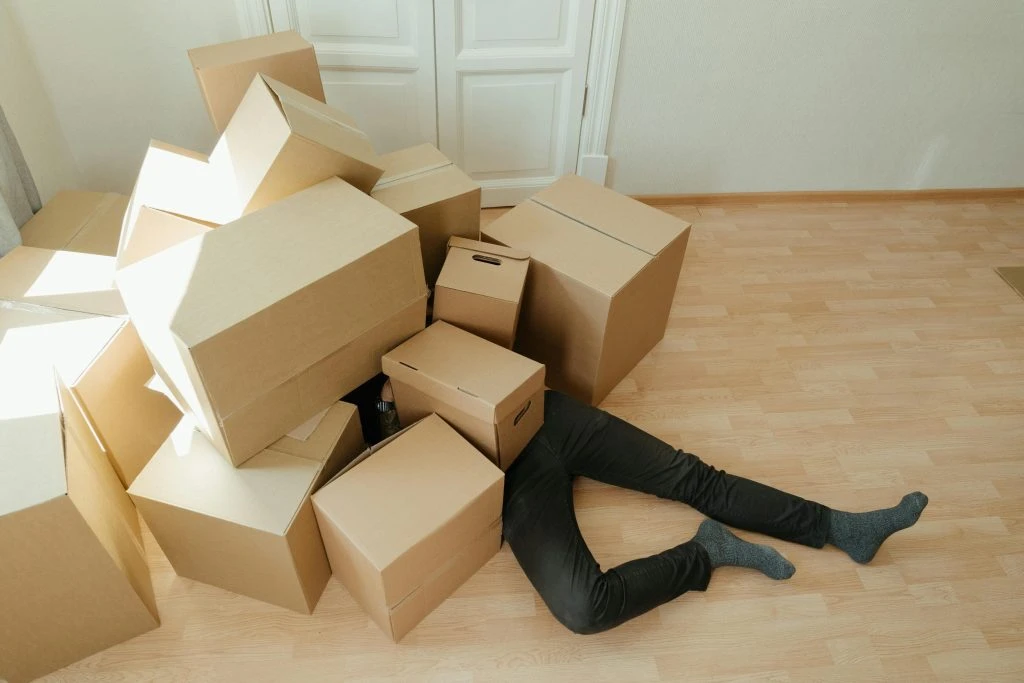 Person lying on floor buried under a pile of boxes.